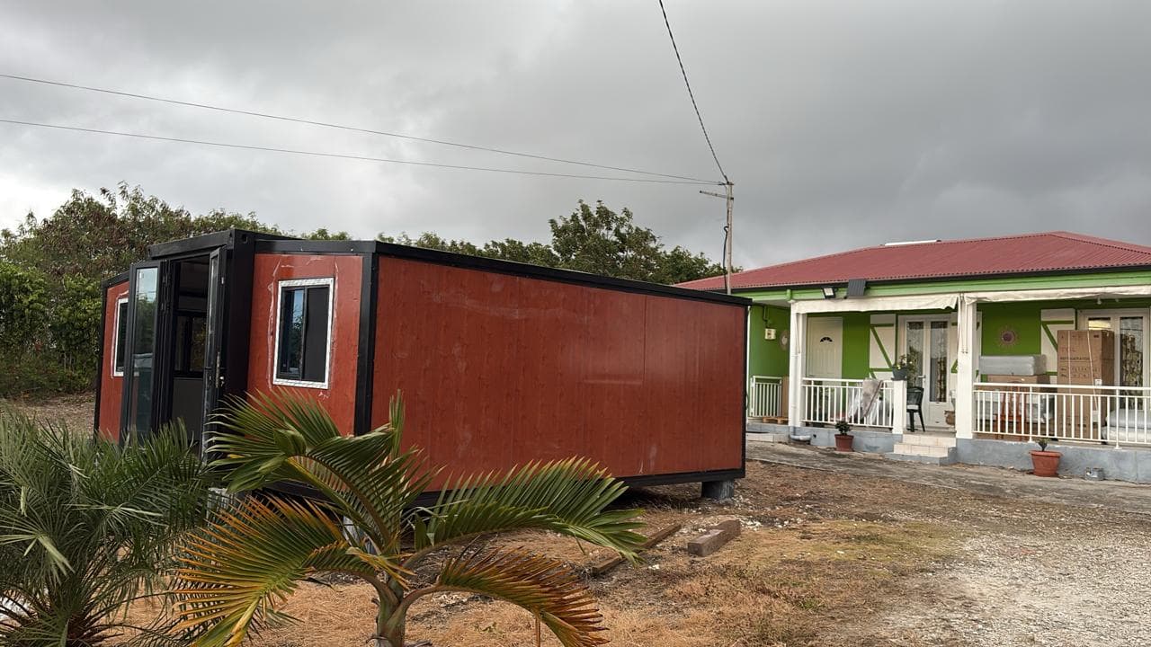 Delivered red expandable home unit sited beside a green house on a rural property.